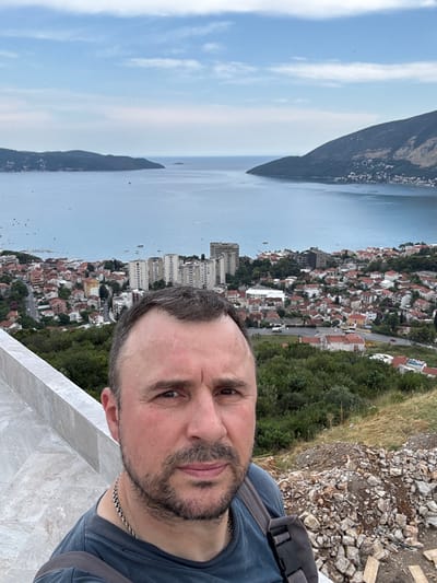 This media file is a selfie taken from a high vantage point, overlooking a coastal town and the sea. The primary subject is a man with short, dark hair, a beard, and a grey t-shirt, wearing a silver necklace. He is looking towards the camera with a neutral expression.
The background reveals a picturesque scene. Below him, a dense town with red-tiled roofs stretches along the coastline, nestled between a steep, tree-covered mountainside and a large body of water, likely a bay or gulf. The architecture appears to be European, with some modern buildings interspersed among older structures. Several boats are visible on the water.
The time of day appears to be daytime, with bright, diffused light, suggesting it might be overcast or late afternoon. The weather seems calm, as indicated by the smooth surface of the water. The location cues, specifically the mountainous terrain meeting the sea and the characteristic architecture, along with the provided location context of Sušćepan, Montenegro, strongly suggest this is a view of the Bay of Kotor. The terrain is rocky and sparsely vegetated in the foreground where the man is standing, possibly indicating a viewpoint or a recently developed area.
There is no visible text in the image.