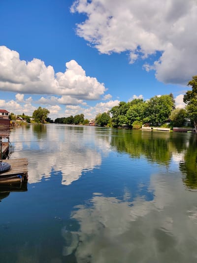 A tranquil river scene unfolds under a bright, partly cloudy sky. The water, calm and reflective, mirrors the dramatic white cumulus clouds and the vibrant blue of the sky. Along the riverbanks, lush green trees create a dense, verdant line, interspersed with glimpses of houses and structures, suggesting a residential area or a village. A wooden pier extends into the water from the left foreground, with a spare tire resting on it, hinting at casual activity or perhaps a mooring point. The scene evokes a sense of peace and a warm, pleasant day, likely late morning or early afternoon, given the strong sunlight casting reflections on the water. The overall impression is one of natural beauty and quiet, rural life.