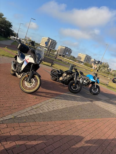 Three motorcycles are parked on a brick-patterned sidewalk in Hook of Holland, Netherlands. In the foreground, a white Honda adventure motorcycle with a black top box faces the viewer. To its right, a black cruiser motorcycle is parked next to a bright blue motorcycle. In the background, modern apartment buildings line a street with trees and streetlights. The sky is blue with scattered white clouds. The scene appears to be captured during daylight hours, likely in the late afternoon due to the long shadows cast by the motorcycles and objects in the foreground.