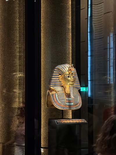 This media file captures a close-up view of an ancient Egyptian sarcophagus, likely a funerary mask of a pharaoh, displayed in a museum setting. The sarcophagus is predominantly gold, adorned with intricate blue and striped patterns, and features detailed facial features, including eyes and a headdress. The object is positioned on a textured, beige surface that is illuminated by warm, directional lighting, creating a spotlight effect. Behind the sarcophagus, a dark, reflective surface suggests a glass enclosure or display case, which also reflects ambient lighting and a distant green exit sign with an upward arrow. The background is dimly lit, with subtle horizontal lines that might indicate architectural elements or further display cases. The overall atmosphere is one of reverence and historical preservation, with the lighting designed to highlight the artifact. The image appears to be taken indoors, likely during daylight hours, given the artificial lighting. There are no explicit people visible as subjects, though reflections in the glass suggest the presence of observers. The location is identified as Kafrat Nassar, Egypt, implying this is an artifact from that region, displayed in a museum.