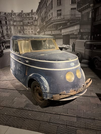 An old, weathered dark blue delivery van or small truck is the central subject of this indoor display. The vintage vehicle shows significant signs of age, with faded and chipped paint, dusty surfaces, and rusty elements. Its front features two large, round, yellowish headlights. A rusty strap or plate is affixed to the front, displaying the numbers "33590". A small, circular emblem, possibly a manufacturer's logo like Citroën, is visible above the headlights. The vehicle's windows are aged and discolored, and a worn tire is visible on the right side.
Behind the car, a large black and white mural depicts a bustling street scene from a past era. The backdrop shows multi-story buildings lining a street, with indistinct figures of people and other vintage vehicles. Prominently visible on awnings or signs are the words "J. DAMOY" and "Julien Damoy," indicating a commercial establishment, likely French given the historical context of the name. A partial sign also reads "TELEP."
The car is presented on a dark-colored floor with a repeating diamond-plate or textured pattern, crossed by thin vertical lines, suggesting a museum or exhibition setting. The overall scene is a static display, highlighting the historical vehicle against a period-appropriate urban backdrop. The lighting is artificial, accentuating the exhibit. The image captures a historical exhibit, likely located in Wassenaar, Netherlands.