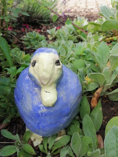 A close-up, eye-level, outdoor shot shows a blue ceramic bird statue with a light yellow face, beak, and feet. The statue is positioned among green foliage and brown soil, suggesting a garden setting. The background consists of more greenery and a blurry, paved area. The lighting is natural, and the focus is sharp on the statue. The image appears to have been taken in Grünwald, Germany.