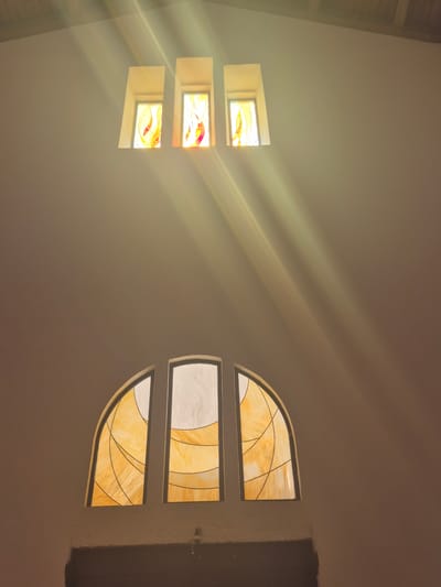 The image depicts the interior of a building with stained glass windows. There are two sets of windows visible. The upper set consists of three rectangular stained glass windows, each featuring abstract designs in red and yellow hues. These windows are positioned high on a light-colored wall, and light beams are streaming through them, creating a dramatic effect. The lower set of windows features three arched stained glass panels with swirling patterns of yellow and orange. These windows are positioned above a dark horizontal structure, possibly a doorway or a ledge. The overall impression is one of serene spirituality, likely in a religious building such as a church or synagogue. The provided context suggests this scene is located in Abu Gosh, Israel, a locality known for its Crusader history and its significant Arab Christian and Muslim populations.