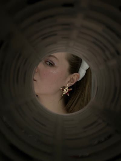 Through a cylindrical opening, a woman's face is visible. She wears a white, fluffy headband and a star-shaped earring adorned with colored gems. The background is out of focus, drawing attention to the subject's face, which is subtly illuminated. The unique perspective creates an intriguing, almost voyeuristic, feel.