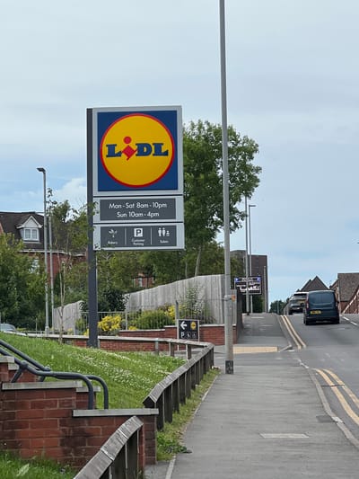 A street sign for Lidl is prominently displayed at a slight angle. The sign features the Lidl logo, a red diamond within a yellow circle, and text indicating store hours: "Mon - Sat 8am - 10pm" and "Sun 10am - 4pm". Below this, there are icons for "Bakery," "Customer Parking," and "Toilets." The sign is attached to a grey pole. To the left of the sign, a brick wall and a grassy slope with small white flowers are visible. Behind the wall, a building with a red-brick exterior and white trim on the roof is partially visible. A street lamp stands to the left of the sign.
The scene continues to a paved road with double yellow lines, indicating no parking. Two vehicles are driving away from the camera up a slight incline: a silver SUV and a dark blue van. Further up the road, a smaller sign is visible on a pole, which appears to direct to parking and cultural attractions. Trees and buildings line both sides of the road. The sky is overcast with light grey clouds. The overall environment appears to be a suburban or commercial area.