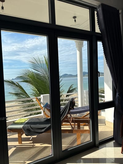A view through a large window shows a balcony with two pieces of furniture and a beautiful ocean and beach view in Song Cau, Vietnam. On the balcony, a wooden lounge chair is covered with a black blanket and a green towel. Behind it, a hammock is suspended between two wooden frames. The balcony overlooks a white railing, a sandy beach, and the turquoise ocean. Palm fronds from a nearby tree are visible in the foreground. The sky is blue with scattered white clouds. A white pillar, part of the building's architecture, is also visible. The scene suggests a serene and relaxing atmosphere, typical of a tropical vacation destination.