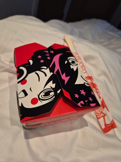 A close-up shot depicts a closed red cardboard takeout container with an anime-style girl's face graphic in black, white, and pink. Red circles are on the girl's cheeks. A pair of wooden chopsticks lies on top of the container, partially obscured by it. The container and chopsticks rest on a white bed sheet with visible folds and creases. The image was taken in Cardiff, United Kingdom.