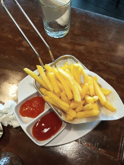 A basket of golden-yellow French fries sits atop a white plate with two small sauce compartments filled with vibrant red ketchup. The fries rest on a white paper liner within the metal basket. A clear drinking glass with a paper napkin inside is visible in the background. The scene unfolds on a dark, textured wooden surface.