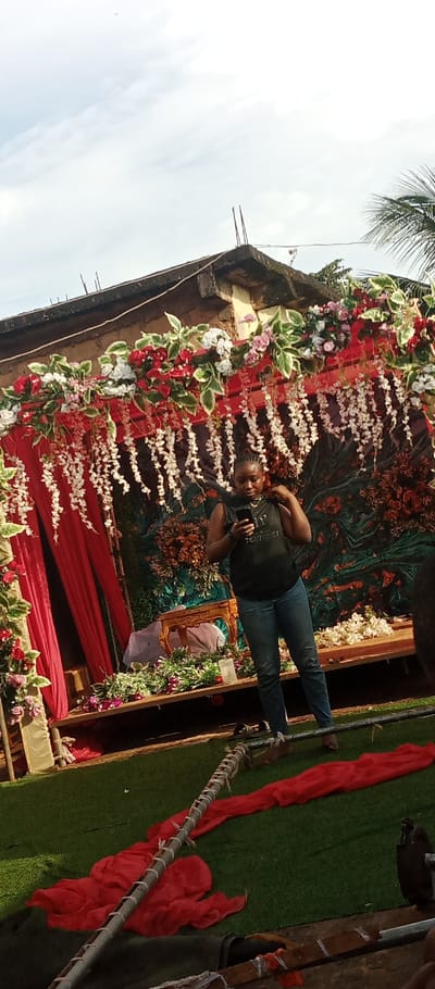 An outdoor scene in Udi, Nigeria, displays a decorated event setup. A dark-skinned female individual stands centrally in the mid-ground, facing slightly left. She is wearing a dark sleeveless top with light text on the chest, blue jeans, and appears barefoot. She holds a dark smartphone in both hands, looking down at the screen, and her right hand is near her left shoulder strap.
Behind her is a raised platform serving as a stage or ceremonial archway. The top section of this structure is adorned with artificial flowers and foliage, primarily red, white, pink, and green, with multiple vertical white floral garlands hanging down. Red drapes are visible on the left side of the background structure. The backdrop behind the individual features dark green, red, and brown textured patterns. On the platform, to the left of the individual, an ornate gold-colored table is visible, along with various floral arrangements and a white cylindrical object.
Above the decorated structure, a section of a concrete building with exposed rebar is visible under a light, cloudy sky. A palm frond is partially visible on the far right side of the upper frame. The foreground consists of artificial green turf, upon which several pieces of loose red fabric are scattered. Two light-colored poles, possibly wrapped in material, lie diagonally across the lower portion of the turf.