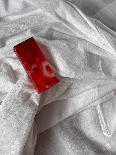 The scene, located in Barabinsk, Russia, depicts a single rectangular bar of translucent red material resting diagonally on a wrinkled white fabric surface. The red bar, approximately 10-12 cm in length, contains numerous small, dark, irregularly shaped specks distributed throughout, resembling seeds, along with several amorphous opaque white swirls. The material exhibits a smooth, glossy surface texture. The white fabric background is heavily creased and folded, creating varying levels of shadow and highlight across its textured surface. No individuals, actions, or additional objects are visible within the frame.