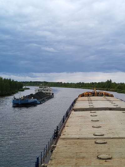 An overcast day in Ozernoe Ust'e, Russia, features a wide, calm river with two barges. In the distance, a blue barge, loaded with dark material resembling coal, navigates the waterway. Its superstructure is white, and it has a small red buoy to its left. In the foreground, the deck of a larger, lighter-colored barge stretches across the frame, revealing numerous circular hatches and yellow safety railings. The banks of the river are lined with dense green trees, under a sky filled with heavy, gray clouds.
