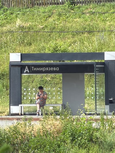 An elderly woman sits alone on a bench at a bus stop. She wears a colorful floral-print dress and holds a smartphone in her hands. The bus stop has a modern design with metal framing and glass panels. The background consists of a grassy slope with a wooden fence at the top, suggesting a semi-urban or suburban setting. The name of the bus stop, "Тимирязева" (Timiryazeva), is visible on the sign. The overall scene is one of quiet waiting in an outdoor environment.