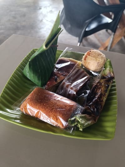 A green, ridged plate holds several Indonesian snacks. On the left, a triangular snack is wrapped in a green banana leaf, secured with a small twig. To its right, a rectangular snack is encased in clear plastic, appearing to be a sweet cake with a slightly granular surface. Next to this, a dark, jelly-like snack is also in clear plastic packaging. Above these, another snack in clear plastic shows a brown, layered confection. To the far right, a small, round, light-colored snack, possibly a ball of sweetened rice or coconut, sits on a banana leaf that's slightly charred and wrapped in plastic. The plate is placed on a plain, light-colored surface, and in the background, a dark chair with wooden legs is visible, suggesting an indoor or semi-outdoor setting, possibly a warung or cafe in Cotgadong, Indonesia. The overall lighting is natural, casting soft shadows.