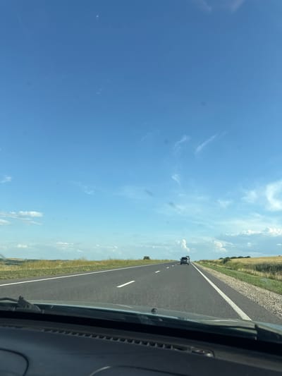 The image depicts a view from inside a vehicle traveling on a paved road in Sagaidac, Moldova. The road stretches into the distance, with a white line marking the edge of the lane. The sky is a vibrant blue with scattered white clouds. To the sides of the road, there are grassy fields and some trees. Two vehicles are visible on the road ahead, traveling in the same direction. The foreground shows the dashboard and windshield wipers of the vehicle from which the image was taken. The overall scene suggests a sunny day and a rural landscape.