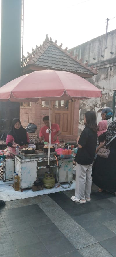 In the city of Kauman, Indonesia, a food stall operates under a large, faded pink umbrella. The stall features two cooking stations, one manned by a woman in a black hijab and pink patterned clothing, and the other by a man in a red shirt. Both are engaged in food preparation. Behind them, a traditional-style building with a tiled roof and decorative elements suggests a historical or cultural area. Several people are gathered around the stall, including customers and what appear to be staff members. The ground is paved with dark gray and lighter gray square tiles. Various items are visible at the stall, such as bottles of oil and sauces, cooking utensils, and a green gas cylinder. The overall scene depicts a typical street food vendor environment.