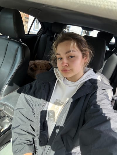 A young woman with light brown hair sits in a car wearing a white hoodie and a dark jacket. A brown poodle sits in the backseat beside her. She looks at the camera with a slight smile. The car's interior is black leather. Outside the car window, one can see buildings and trees. The image is rotated counterclockwise, with the car door on the left.