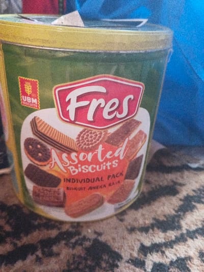 This image depicts a cylindrical container of "Fres Assorted Biscuits" in Dogsenang Gorphoog, Bhutan. The container is predominantly green with a yellow rim and displays a red label featuring "Fres" in white script. Below "Fres," the text "Assorted Biscuits Individual Pack" and "BISKUIT ANEKA RASA" are visible, surrounded by illustrations of various biscuit types including wafers, round cookies, and rectangular biscuits. A "UBM BISCUITS" logo with a wheat stalk icon is present on the upper left side of the label. The container rests on a dark brown and black patterned surface, likely a rug or carpet. A bright blue, indistinct object is partially visible in the blurred background to the upper right of the container. No individuals are present in the scene, and no specific actions or interactions are depicted.