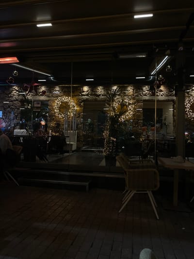 The image, oriented vertically, depicts a nighttime view of an outdoor or semi-enclosed area of a cafe or restaurant in Chaidari, Greece. On the right side of the frame, a dark brick pavement features a stack of light-colored, possibly wicker or rattan, chairs next to a table, suggesting the area might be closed or prepared for outdoor dining.
A clear glass partition runs horizontally through the center of the image, separating the foreground from a background area. Beyond the glass, a textured stone wall is adorned with several circular wreaths and scattered garlands of warm-toned string lights, creating a festive and cozy atmosphere. Some green plants are visible among the illuminated decorations.
Above the glass partition, a dark, slatted ceiling structure is fitted with modern rectangular LED strip lights. Through the glass, glimpses of other tables and chairs can be seen, and at least two individuals are present; one person is partially visible in the bottom right corner of the frame, seated with their back towards the viewer. The entire scene is illuminated by artificial lighting, indicating it is evening or night. A small sign resembling the American Express logo (a blue square with a white symbol/text) is discernible on the stone wall behind the glass partition.