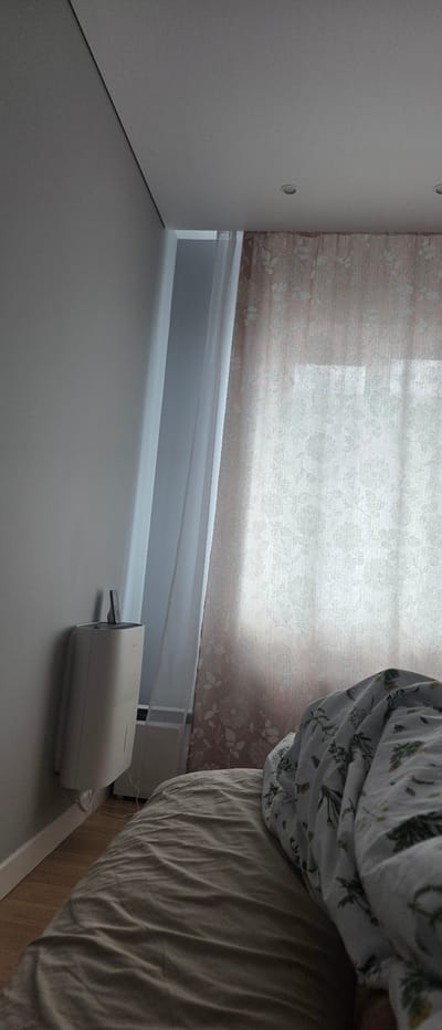 The image depicts an indoor scene, likely a bedroom, with a bed in the foreground covered by a duvet featuring a floral pattern. To the left of the bed, a white dehumidifier is mounted on a light gray wall. Behind the dehumidifier, sheer white curtains are visible, and beyond them, patterned pink curtains cover a window. The room has a neutral color palette with light walls and wooden flooring. The lighting suggests it is daytime, with natural light filtering through the curtains. The image does not contain any individuals or specific actions. There is no information that would allow for locating this scene in Belyaninovo, Russia.