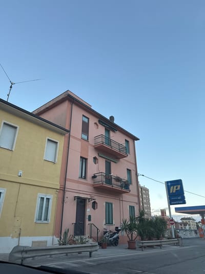This image depicts a street scene at Via S. Giovanni, 44, Grottammare, Italy.

**Subjects and Objects:**
The primary subjects are two adjacent buildings and a partial view of a gas station. On the left, a two-story residential building with a pale yellow facade features two windows with closed white external roller shutters on the second floor and one on the first floor. A television antenna is mounted on its roofline, extending into the clear blue sky.

To its right stands a four-story residential building, painted in a faded pink hue with darker vertical pink accent strips. The ground floor entrance features a dark brown door, accessible via two dark-colored steps with a metal handrail, and is protected by a small dark canopy. Two potted agave plants flank the entrance. This building has multiple windows with dark frames; some have closed dark green louvered shutters. Two balconies with black metal railings are present on the second and third floors; the lower balcony contains several potted plants. A silver scooter (motorcycle) with a black seat and top case is parked on the sidewalk near the building's entrance.

To the right of the pink building, a blue "IP GRUPPO api snack bar" sign is visible, indicating the presence of a gas station, partially obscured by power lines. A white gas station canopy is visible beneath the sign. In the distant background, a taller, multi-story beige building and other smaller structures are discernible.

**Environment and Actions:**
The sky is clear and light blue, indicating daytime conditions. No individuals are visible in the scene. A silver guardrail is present in the immediate foreground, suggesting a roadside perspective, possibly from a vehicle. The ground consists of asphalt road and a paved sidewalk area. Potted plants are arranged along the sidewalk in front of the pink building and extending towards the gas station.