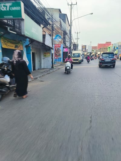The image shows a street scene in Bojongsoang, Indonesia, taken from a moving vehicle. The camera is facing forward. A person wearing a black abaya walks across the street from right to left. They are walking away from the camera. There is a white scooter in the middle of the road with a person wearing a pink shirt and a white helmet riding it towards the camera. In the background, there is a truck parked on the right side of the road, and there are a few other vehicles in the distance. There are power lines and electrical wires running along the side of the road. The buildings lining the street have signs, including one that says "J&T Cargo." The road is paved and appears to be in good condition. The sky is clear and blue, with a few white clouds. There is a streetlight in the middle of the road. The location in Bojongsoang, Indonesia, based on coordinates (latitude: -6.982, longitude: 107.634) is a residential area with a mix of businesses and homes. The image is taken in a sunny, midday condition.