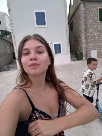 In Primošten, Croatia, a young woman with long brown hair is taking a selfie outdoors in what appears to be a pedestrianized area with stone buildings. She is wearing a dark-colored top with a sequined pattern and a delicate necklace. To her right, a young boy in a white shirt with a palm tree print is looking away from the camera while holding onto a blue scooter. The background features traditional stone architecture with blue shutters and a green garbage bin. The lighting suggests it is either early morning or late afternoon.
