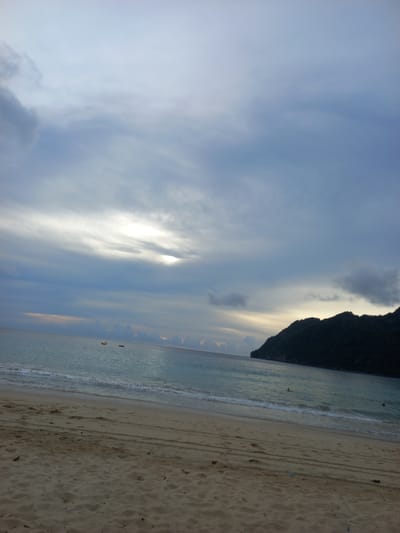 The image depicts a beach scene at Meunasahbale, Indonesia. The sky is overcast with dramatic clouds, featuring a bright break in the clouds where the sun is partially visible, casting a soft light on the water. The ocean is calm with gentle waves lapping at the sandy shore. In the distance, a dark, tree-covered hill or mountain rises from the water on the right side of the frame. Two small yellow objects, possibly buoys or small boats, are visible on the water. One person is swimming in the ocean on the right side of the frame. The sandy beach in the foreground shows tire tracks, suggesting vehicle activity. The overall atmosphere is serene and somewhat subdued due to the cloudy weather.