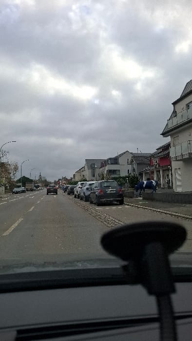 The image displays a street scene in Frisange, Luxembourg, captured from inside a moving vehicle facing generally southeast on Rue Robert Schuman (CR153). The sky is overcast with a uniform grey cloud cover.
The foreground is partially obscured by the vehicle's dashboard and a black suction cup mount on the lower right windshield. The asphalt road features two lanes, one for each direction of travel, marked by white lines. The vehicle is positioned in the right lane. Approximately eight passenger cars are visible parked parallel to the curb on the right side of the street, including a dark-colored Fiat 500 in the mid-ground. An additional dark-colored vehicle is observed traveling ahead in the same lane. In the opposing lane, a light-colored truck or van is visible in the distance.
On the right side of the road, a paved sidewalk abuts a row of multi-story buildings of varying modern architectural styles. The closest building exhibits a light-colored facade with red accents and glass-fronted balconies. Positioned on a low wall or planter on the sidewalk in front of one of these buildings is a life-sized, blue and white painted cow statue, facing towards the street. Further down the street on the left, streetlights with curved arms are visible. In the distant background, the distinct spire of the Église Saint-Martin de Frisange is clearly identifiable. No individuals are visible in the scene. Scattered trees, many with bare branches, line parts of the street.