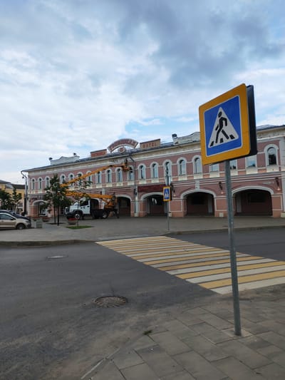 An exterior, medium shot captures a pedestrian crossing in front of a pink, historic hotel in Vasil'ki, Russia. The hotel, featuring arched doorways and windows, is adorned with a sign that reads "Gostinitsa" and "Hotel". Above the hotel, a crane with a worker is visible, suggesting ongoing maintenance or construction. The sky is partly cloudy. The street is paved, and a yellow and white striped pedestrian crossing is prominently displayed. A blue and yellow pedestrian crossing sign stands to the right of the frame. Cars are parked on the left side of the image, and a manhole cover is visible on the street.