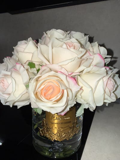 An image, rotated 90 degrees counter-clockwise, displays a bouquet of artificial roses arranged in a clear glass bottle. The bottle is lying horizontally on a dark, reflective surface, with a textured, grayish wall or background behind the roses.

The roses are primarily white or very pale cream, with many individual petals featuring delicate pink edges. Some roses reveal a soft peach or light pink hue in their densely packed centers, suggesting their fabric material. A few small green leaves are interspersed among the blossoms.

The glass bottle, acting as a vase, features an ornate, gold-colored label with intricate detailing. Visible text on the label includes "Colette Noire" and "PARFUM DE MAISON," indicating it is likely a decorative home fragrance bottle from a brand named "Maison de Colette Noire" or similar. The lighting appears artificial, casting soft shadows and highlighting the textures of the roses and the metallic sheen of the label. There are no people or specific activities depicted. The setting is indoors in Zvezditsa, Bulgaria.