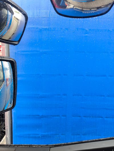 The image shows a view from inside a vehicle, likely a truck, looking out towards a bright blue tarp or curtain. The blue surface is covered in numerous small water droplets, indicating recent rain or a car wash. The texture of the tarp suggests it's a woven material.

Reflections in the vehicle's side mirrors offer glimpses of the surroundings. The left mirror reflects the side of a blue vehicle, possibly another truck, with some stickers or labels visible. The right mirror, positioned higher up, reflects a street or pavement made of interlocking concrete blocks, and a building or structure with a red and white accent. The overall lighting suggests it is daytime.

The scene appears to be at a location in Młyny, Poland, possibly a rest stop, truck wash, or loading/unloading area. The presence of water droplets and the vehicle context suggest an activity related to transit or vehicle maintenance. No specific emotions are conveyed, and the overall impression is one of an ordinary, functional environment. There is no legible text in the primary view, though faint text might be present on the reflections in the mirrors, which is too blurry to decipher.