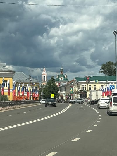 The image shows a street scene in Pereslavl-Zalessky, Russia, on a cloudy day. The view is from a vehicle traveling on a road with double white lines. On the left side of the road, there is a yellow building decorated with numerous Russian flags on flagpoles. In the background, several church domes and spires are visible, characteristic of Russian Orthodox churches, including a prominent bell tower with a white and red design. On the right side of the road, there are other buildings, traffic lights, and several vehicles, including a white truck and a white SUV. The sky is filled with dark, dramatic clouds.
