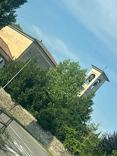 The image shows a view of a building with a bell tower, surrounded by trees and vegetation, with a street in the foreground. The building appears to be made of stone and has a tiled roof. The bell tower is square and has arches with bells inside. A cross is visible at the top of the bell tower. The trees are lush and green, and some appear to be quite old. The street is paved and has white lines marking the lanes. The sky is blue and clear. This image captures a peaceful scene in Pumenengo, Italy, likely in a rural or semi-rural area given the abundance of greenery and the presence of a church bell tower.