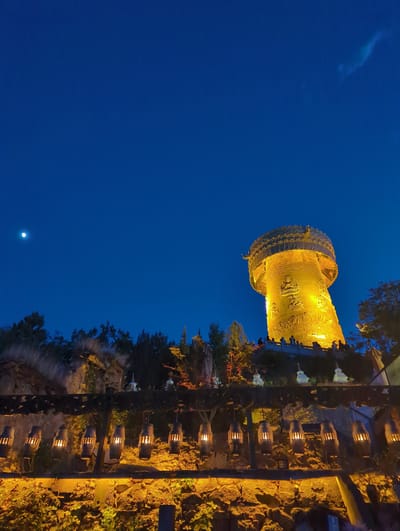 The image shows a nighttime scene in Shangri-La, China, featuring a large, illuminated structure resembling a golden prayer wheel or stupa. The structure is adorned with intricate carvings and appears to be made of metal, possibly gilded. It is lit from within and by external lights, casting a warm, yellow glow against the deep blue night sky.

In the foreground, a rustic pathway is lined with numerous small, lantern-like lights, also emitting a warm, inviting glow. These lanterns are housed in what appear to be wooden casings, some resembling miniature barrels. The pathway is bordered by textured, rocky ground and lush green foliage, with some trees visible in the background. The overall atmosphere is serene and spiritual, characteristic of a temple or cultural site.

The sky is clear and dark, indicating nighttime, and a faint moon or bright star is visible in the lower left quadrant of the image. The rotation of the image suggests it might have been taken with the camera held sideways. The scene evokes a sense of peace and cultural richness. No people are directly visible in the frame, and no text is discernible.