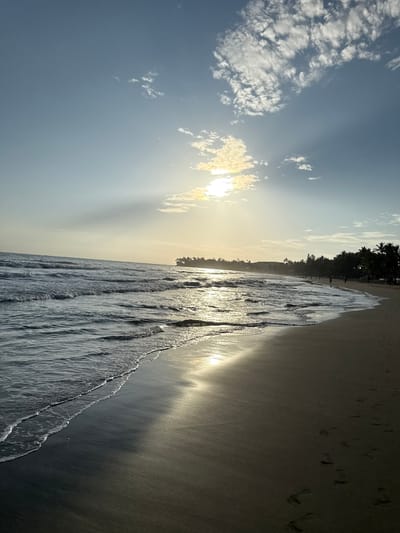 The image depicts a beach scene at Cabarete, Dominican Republic. The sun is low in the sky, shining through a few clouds, casting beams of light. The ocean waves wash onto a sandy beach, reflecting sunlight. Footprints can be seen in the sand. A line of palm trees is visible in the background.