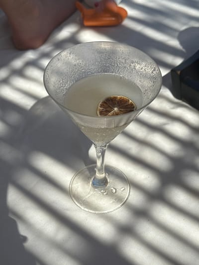 The image depicts a close-up, high-angle view of a clear martini glass containing a pale yellow or clear liquid cocktail, with a single dried citrus slice (likely lime or lemon) floating on the surface. The glass exterior displays significant condensation or frost. It is positioned on a light-colored, possibly white, fabric surface. Strong, distinct parallel shadows, indicative of direct sunlight passing through a slatted overhead structure, are cast across the surface and the base of the glass.

In the upper left background, a bare human foot, likely belonging to the individual whose perspective is captured, is partially visible. In the upper right background, an orange, vaguely boat-shaped or toy-like object and a dark, rectangular object are partially visible and out of focus. No direct actions or interactions are depicted beyond the static arrangement of objects. The environment is consistent with a leisure setting, such as a poolside or patio area at a resort or hotel, within a beach destination in Mexico. The specific city "The Beach, Mexico" does not exist.