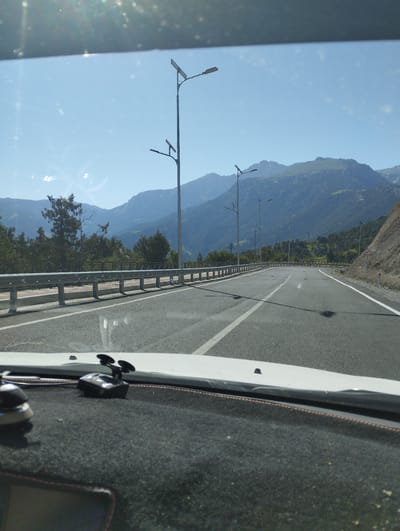 The image depicts a view from the interior of a vehicle driving on a paved road in a mountainous region, likely near Myk, Uzbekistan. The road is asphalt with white lane markings, and there are guardrails along the left side. Streetlights are visible at intervals along the road. In the background, there are large, green-covered mountains under a clear blue sky. The dashboard of the vehicle is visible in the foreground, with a black textured surface and two suction-cup mounted devices. The scene is illuminated by bright sunlight, casting shadows on the road.