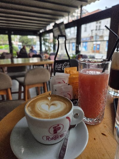 The image depicts a close-up view of a cafe table in Gevgelija, North Macedonia. In the foreground, on a light-colored wooden table, sits a white ceramic cup and saucer. The cup contains a latte adorned with a heart-shaped latte art design and features a red "PORTION" logo. A small silver spoon rests on the saucer. To the right, a clear, textured glass contains a reddish-pink beverage with two black straws. Behind these, a stack of white and orange packets, likely sugar or sweeteners, stands upright next to a black cutout silhouette of a Coca-Cola bottle. The midground shows the interior of a cafe with a dark-beamed ceiling structure and glass walls. Two blurred individuals are seated at a wooden table in the background, one in dark attire and the other in a blue shirt. Through the glass walls, a blurred urban street scene is visible, featuring light-colored buildings with windows and some blue signage under an overcast sky.