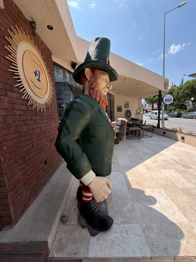 The image shows a statue of a leprechaun holding a beer mug, standing outside a building with a brick facade and a white awning. The statue is life-sized and is dressed in a green jacket, a tall green hat, and striped socks. The building appears to be a cafe or bar, as evidenced by the outdoor seating area and a sign that says "bein SPORTS MAC YAYINI". The scene is set on a sunny day with a clear blue sky and some clouds. In the background, there are cars driving on a street and trees lining the sidewalk. The location is identified as Denizli, Turkey, specifically near the entrance of a establishment that appears to be a sports bar or pub.