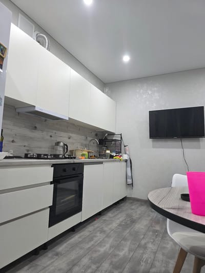 The image depicts a modern, minimalist kitchen in Krymsk, Russia. The scene is well-lit, suggesting it could be daytime. The kitchen features sleek white cabinetry and light grey wood-look flooring. A prominent black television is mounted on a textured grey wall.
On the countertop, a stainless steel kettle and a faucet are visible. There are also various kitchen items and containers, hinting at recent use or preparation. A black oven is integrated into the cabinetry, with its glass door revealing an interior light. The overall impression is one of a clean, organized, and functional living space. There is no visible text in the image.