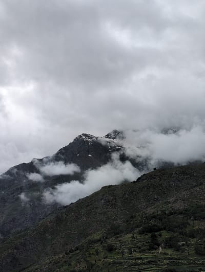 The image depicts a cloudy, overcast day in the mountainous region of Stingri, India. The foreground features a steep, dark, rocky mountainside covered in dense green vegetation, including trees and terraced fields. In the mid-ground, a prominent, rugged mountain peak emerges, partially shrouded by thick, white clouds. Patches of snow are visible on the upper slopes of this peak. The sky above is filled with heavy, gray clouds, creating a dramatic and moody atmosphere. The overall impression is one of natural, untamed wilderness under a somber sky. There are no visible people or man-made structures apart from the terraced agricultural areas on the mountainside.