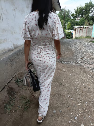 A woman walks away from the camera on a dirt path in Mady, Kyrgyzstan. She is wearing a long, white floral dress with flutter sleeves. She carries two bags, one a woven straw bag and the other a black tote bag, in her left hand. She is also holding a phone in her right hand. The path is lined by a rough concrete wall on the left and a gravelly, uneven surface on the right. In the background, there are trees, a brick wall, and a few buildings. The sky is overcast.