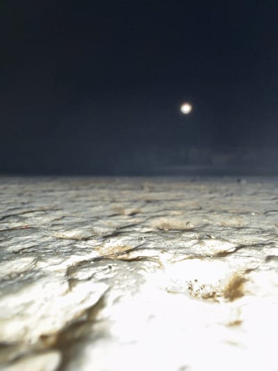 This is a low-angle, close-up shot of a textured, rocky surface, possibly pavement or natural stone, illuminated by a bright light source creating strong highlights and shadows. The surface appears wet or reflective. In the upper background, a dark, hazy sky is visible with a blurry, bright circular object that could be the moon or a distant light. The scene is devoid of any people or specific identifiable landmarks that would suggest the location within the city of Tyulenovo, Bulgaria.