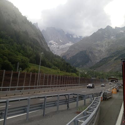 The image depicts a multi-lane highway passing through a mountainous valley, possibly in the vicinity of Courmayeur, Italy, given the presence of a glacier and steep, wooded slopes characteristic of the Alps. The sky is overcast with dense clouds.

On the left side of the highway, a steep mountain slope covered in dense green foliage rises sharply. In the mid-ground, a wooden sound barrier runs parallel to the road. Beyond the sound barrier, the mountain slope continues upwards, revealing rocky terrain and patches of snow further up. A tall, metallic pylon, possibly for power lines or a ski lift, is visible on the slope.

The highway itself has at least three lanes visible in the foreground. Several cars are driving on the highway, including a dark-colored sedan in the left lane and two other cars further ahead. The road is equipped with metal guardrails and lighting poles.

In the distance, the valley opens up to reveal a large, imposing glacier cascading down between rugged, snow-capped mountain peaks. These peaks are partially obscured by clouds. The overall environment is dominated by natural grandeur, with the man-made highway cutting through the dramatic mountainous landscape.

On the right side of the highway, further along the road, a tunnel entrance with an arched structure is visible, and construction or maintenance equipment, including yellow barriers and metal structures, are present near the road edge. The image appears to be taken from a vehicle traveling on the highway.