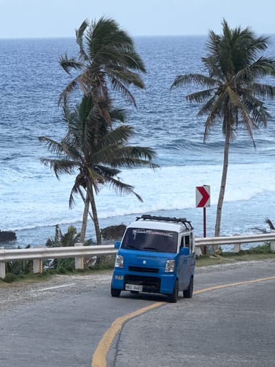The image shows a blue and white vehicle parked on a paved road next to a metal guardrail. The road is marked with a yellow line. Beyond the guardrail, there are palm trees and a view of the ocean with white waves. A road sign with red and white chevrons is visible near the vehicle. The scene is in Burgos, Philippines.