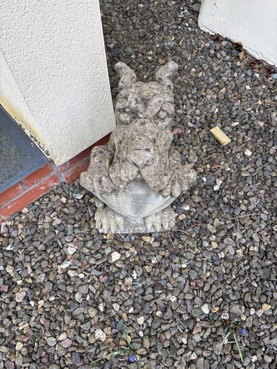 The image depicts a garden setting in Longden, United Kingdom. A stone statue of a turtle sits amidst a bed of grey gravel, bordered by a pale, textured concrete structure and a red-brick edge. A small wooden splinter lies on the gravel near the statue. The scene is viewed from an overhead perspective, focusing on the arrangement of the statue and its surrounding elements.