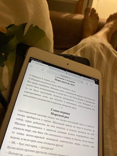 The image shows a close-up view of a tablet screen displaying a book in Russian. The tablet is positioned on a soft, fabric surface, possibly a blanket or sofa, with a hint of a floral pattern visible in the background. The scene appears to be indoors, with warm, ambient lighting. The book title visible on the screen is "Повелитель мух" (The Lord of the Flies). The text also mentions "Перевод. Е. Суриц, 2013" (Translation by E. Surits, 2013). Part of a person's legs, covered by a striped fabric, are visible in the upper right portion of the image, suggesting someone is holding or using the tablet. The time displayed on the tablet is 20:50. No elements in the image suggest a specific location within the city of Kushchovskaya, Russia.