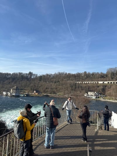 An outdoor, daytime scene at the Rhine Falls in Neuhausen am Rheinfall, Switzerland, depicts six individuals on a paved viewing platform overlooking the Rhine River. The sky is clear blue with multiple white contrails. The river, on the left, exhibits turbulent, foamy white water flowing rightwards, indicating a strong current or waterfall.
From left to right on the platform:
1. A person in a yellow puffer jacket and grey backpack is partially visible, facing right.
2. A person in a dark jacket and black cap holds a dark electronic device, aiming it towards the water.
3. A person in a dark puffer jacket, light blue jeans, a light beanie, and a brown shoulder bag holds a black smartphone horizontally, apparently photographing the river.
4. A male in a white puffer jacket, blue jeans, and sunglasses leans on the metal railing, smiling towards the camera, posing for a photograph.
5. A female in a black puffer jacket and wide-leg black pants stands facing the river or the posing male.
6. A person in a dark jacket, dark beanie, and backpack holds a smartphone horizontally, directing it towards the river.
The platform is made of grey interlocking pavers, with a black metal railing separating the viewing area from the river. Strong sunlight from the right casts prominent shadows of the railing and individuals on the pavement.
Across the river, a steep hill covered in bare deciduous trees rises. Structures on the opposite bank include a large, light-colored building directly adjacent to the water on the far left, a smaller building with an orange roof further right, and another light-colored building with a flat roof near the water on the right side of the river. A long, arched stone viaduct traverses the upper part of the forested hillside in the distance.