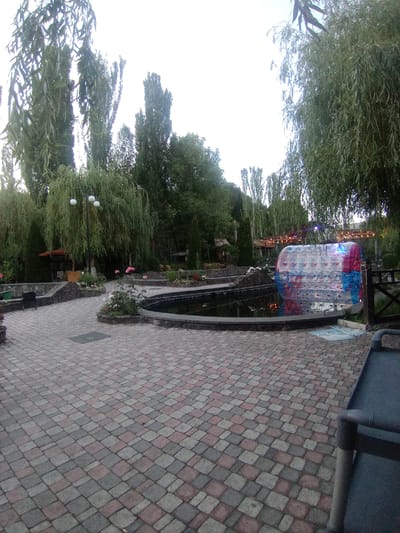In the city of Katnaghbyur, Armenia, a paved area with a patterned brick walkway is visible. Tall trees with dense foliage line the perimeter of the scene, creating a natural canopy. In the center, a pond with a dark water surface is surrounded by a stone wall. Floating in the pond is a large, transparent inflatable sphere, illuminated by blue and pink lights. The sphere appears to be a recreational attraction. Beyond the pond, a wooden structure with overhead lights is visible, suggesting an entertainment or dining area. In the foreground, a dark-colored lounge chair is positioned on the walkway. The sky above is overcast, with a bright, washed-out appearance.