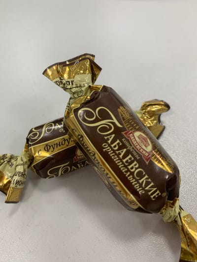 Two individually wrapped chocolate candies are displayed on a white, textured surface. The wrappers are a metallic gold and dark brown, with Cyrillic lettering and a graphic of what appears to be an architectural building. The candies are oriented at an angle, with one partially obscuring the other. The background is a plain, light gray. No location in Urtaaul, Uzbekistan can be determined from the image.